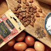 A wooden board showcases a bag of GW LUV HEARTS, loose heart-shaped dog treats, six brown eggs, cloves, a blue bowl of flour, and a rolling pin—perfect for baking or making treats.