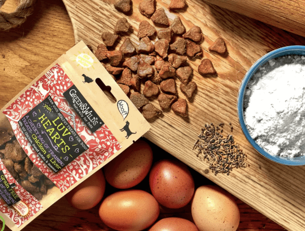 A wooden board showcases a bag of GW LUV HEARTS, loose heart-shaped dog treats, six brown eggs, cloves, a blue bowl of flour, and a rolling pin—perfect for baking or making treats.