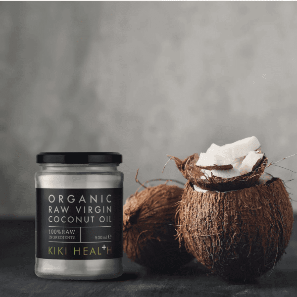 Screenshot 2025-05-24 at 13.57.46 A jar of Kiki Organic Raw Virgin Coconut Oil - 100% Raw is placed next to two brown coconuts, one split open to show the white coconut flesh, set against a gray background.