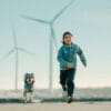 A young child in a denim jacket runs joyfully on a sandy path beside a small dog wearing the Truelove Easy Fit Quick-Release Pet Harness with Dual D-Rings, with large wind turbines and a clear sky in the background.