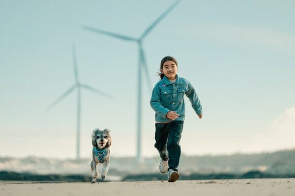 A young child in a denim jacket runs joyfully on a sandy path beside a small dog wearing the Truelove Easy Fit Quick-Release Pet Harness with Dual D-Rings, with large wind turbines and a clear sky in the background.