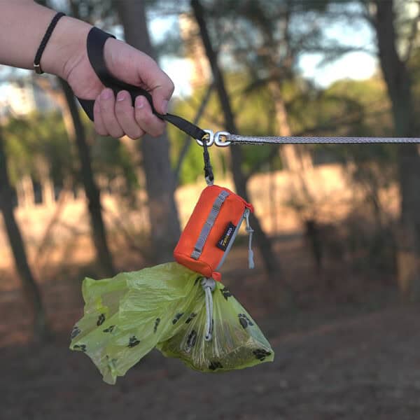A hand holds a dog leash with an orange waste bag dispenser and green bag, outdoors—perfect for walks or trips with your Truelove TLB2012 Waterproof Multi-Use Pet Carrier made from DuPont material.