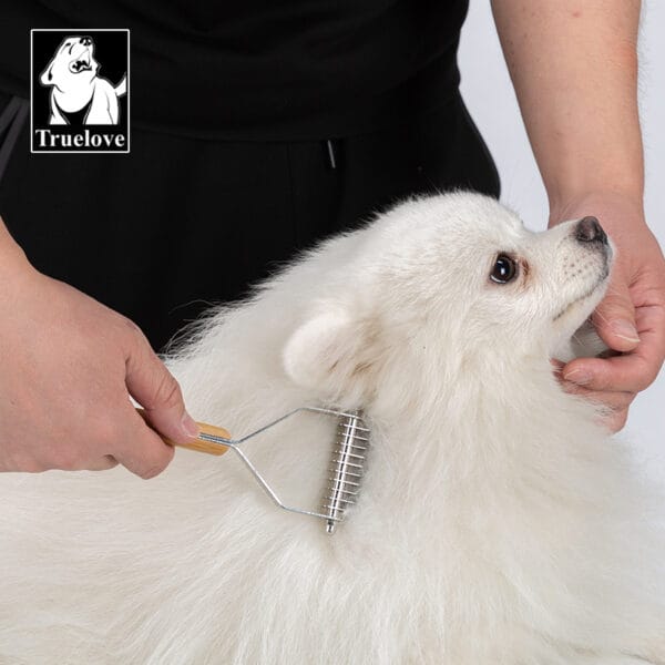 A person gently brushes a fluffy white dog with the Truelove Bamboo Molting Hair Comb, which has a bamboo handle. The calm dog is beside a "Truelove" logo with a dog's face in the top left corner.