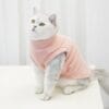 A white and gray cat with green eyes sits on a white surface, wearing the Fleece Dog Sweater | Warm Winter Vest for Dogs & Cats in soft light pink, against a plain white curtain background.