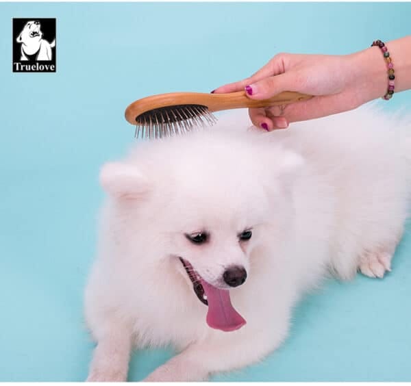 A fluffy white dog with its tongue out is brushed by a person using the Truelove Bamboo Pet Grooming Comb – Premium Brush, against a light blue background. The Truelove logo appears in the top left corner.