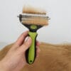 A hand holds the 2 in 1 Pet Grooming Comb - Mats & Loose Hair Removal Brush with light brown fur in its bristles, matching the fur below—likely from a dog.