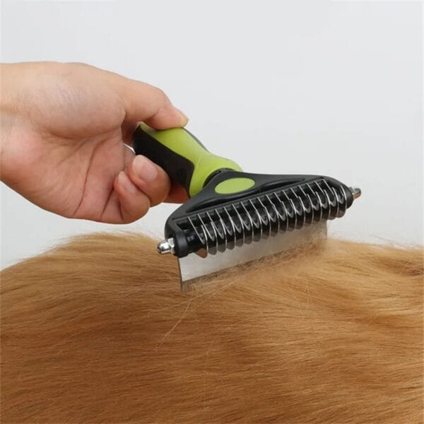 A hand uses a green and black pet grooming brush to comb the fur of a light brown animal, likely a dog, against a plain white background.
