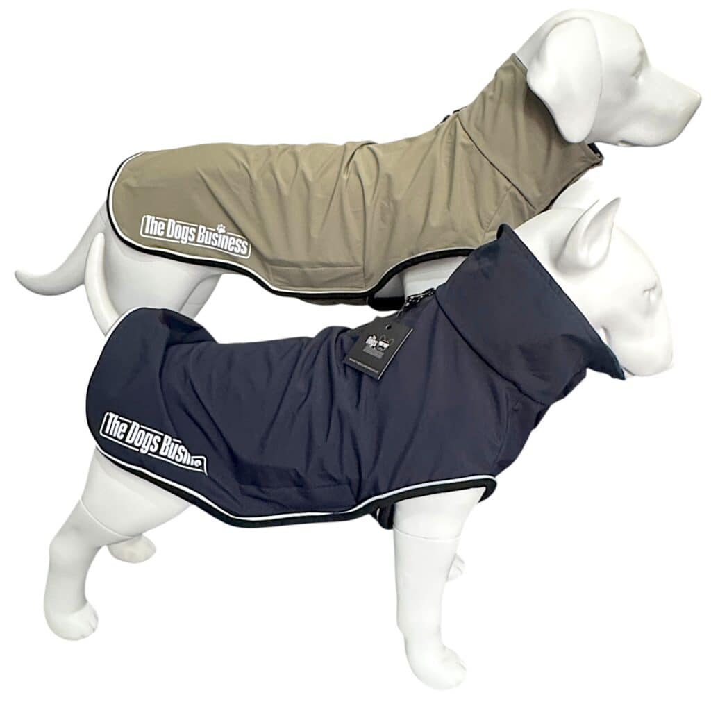 Two white dog mannequins wear TDB light Jackets—one tan, one dark blue—each labelled "The Dogs Business." Both coats have high collars, waterproof fleece lining, and white piping along the edges.