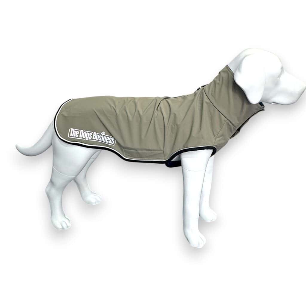 A white dog mannequin wearing a light brown, waterproof dog coat with black trim and the text "The Dogs Business" printed on the side. The background is plain white.