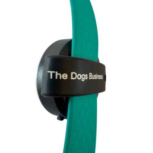 A close-up of The Dogs Business Safety Light, a black circular device with a teal band labelled “The Dogs Business” in white text, shown against a plain white background.