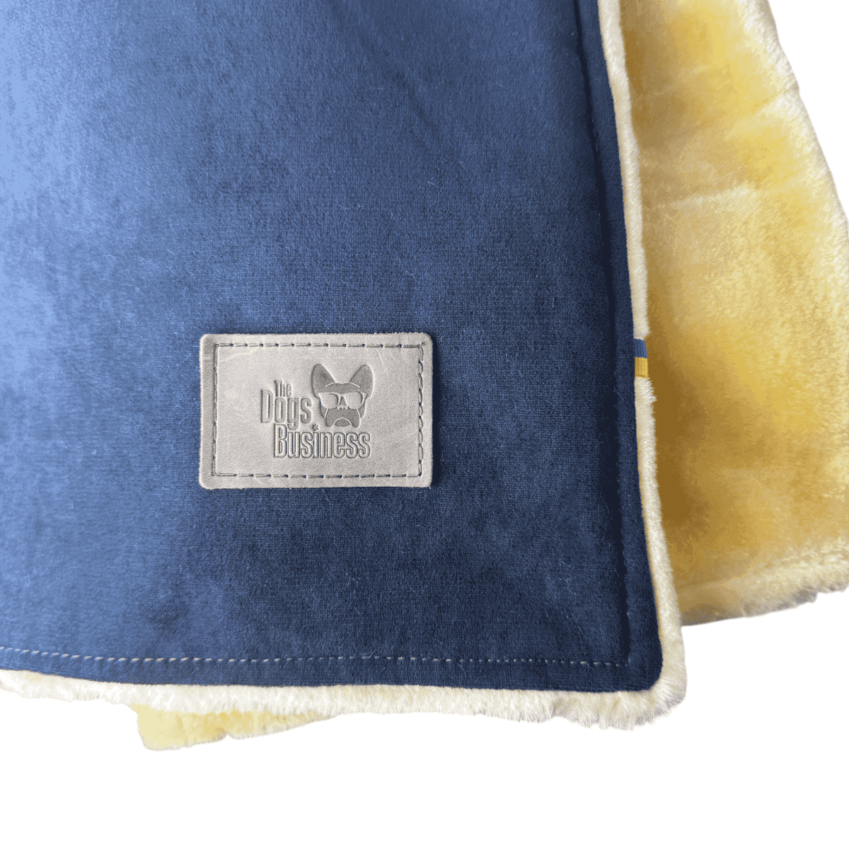 Velour snuggle blanket in blue with a yellow faux-sheepskin lining and a rectangular patch featuring a logo of a French bulldog wearing sunglasses and the text “The Dog’s Business”.