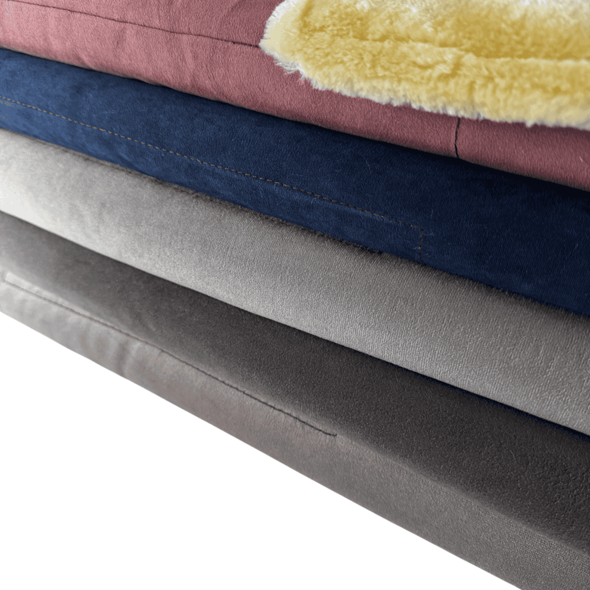 A stack of four folded Velour Snuggle Blankets in dusty pink, navy blue, light grey, and charcoal grey, with a corner of a yellow sheepskin-style blanket folded over the top.